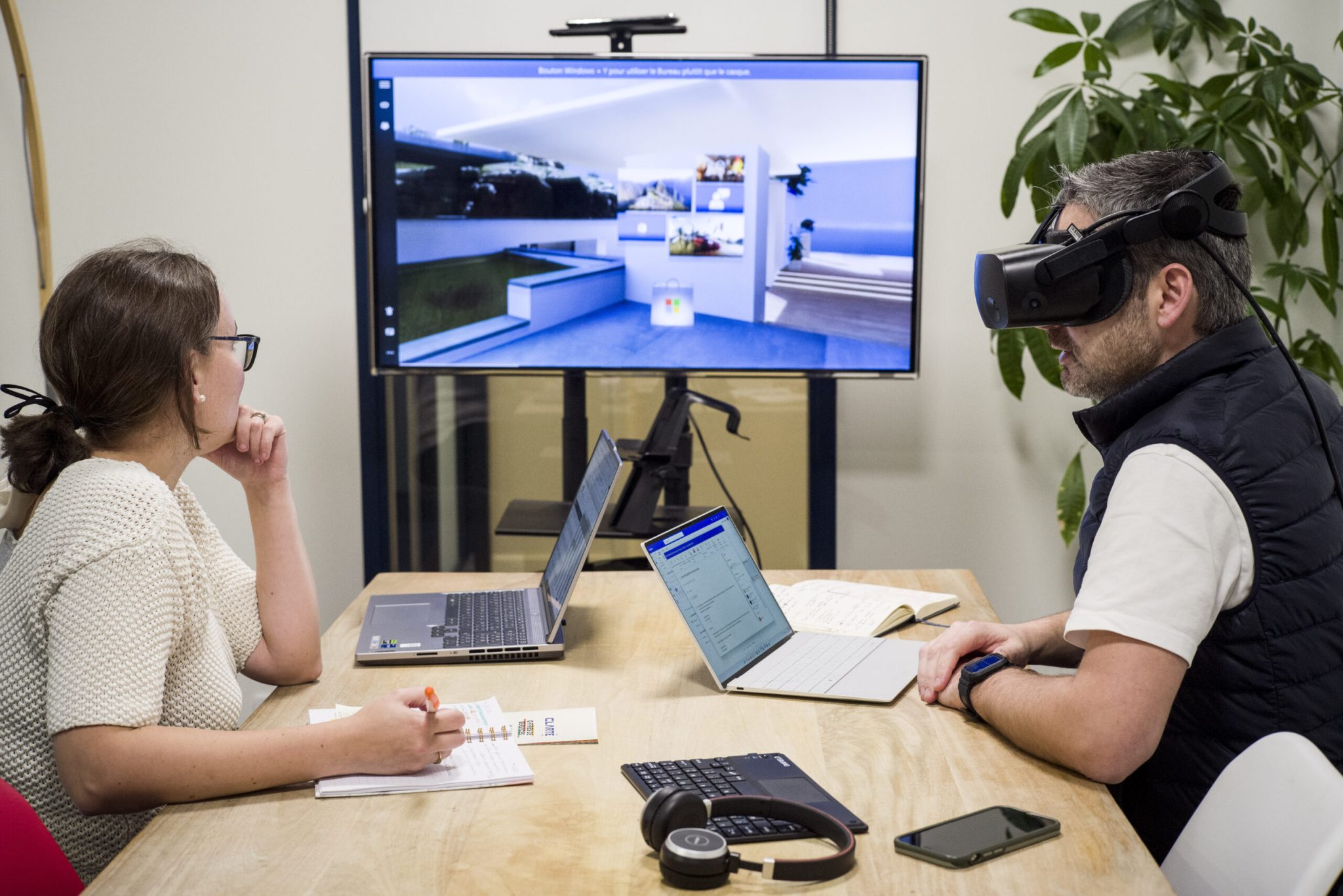 Deux personnes en réunion, une personne avec un casque de réalité virtuelle, et l'autre personne regardant ce que la personne voit dans le casque à l'aide d'une télévision